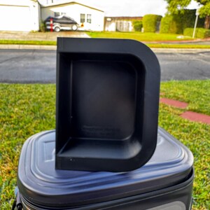 Puede incluir: Un contenedor de almacenamiento de pl&aacute;stico negro en forma de D se encuentra sobre una nevera blanda. El contenedor tiene una base rectangular y una parte trasera curva y elevada. La nevera es negra y gris con la marca "RTIC" visible.