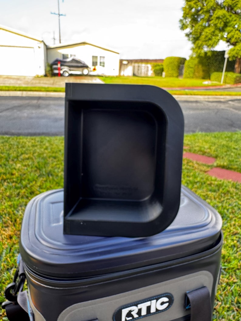 Puede incluir: Un contenedor de almacenamiento de pl&aacute;stico negro en forma de D se encuentra sobre una nevera blanda. El contenedor tiene una base rectangular y una parte trasera curva y elevada. La nevera es negra y gris con la marca "RTIC" visible.