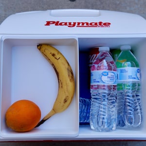 May include: A white Playmate cooler with red lettering, divided into two compartments. One side holds a banana and an orange. The other side contains bottled water and soda. The cooler is open, revealing its contents.