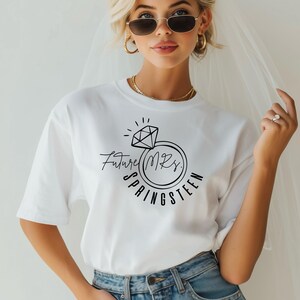 May include: White t-shirt with a black graphic of a diamond ring and the text "Future Mrs. Springsteen". The shirt is a casual style, perfect for a bridal event.