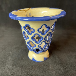 May include: A ceramic vase with a cream-colored body and blue trim. The vase features a series of blue, four-petal cutouts around the body. The rim and base are also trimmed in blue, creating a decorative piece.