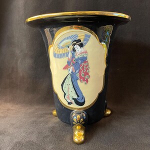 May include: Black vase with gold trim and three gold feet. The vase features a painted image of a woman holding an umbrella. The woman is wearing a blue kimono and is set against a cream-colored background.
