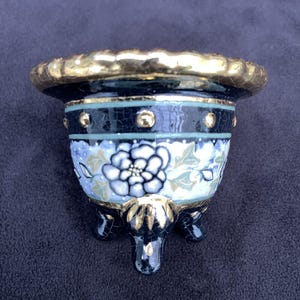 Può includere: Piccolo vaso decorativo in ceramica con bordo dorato e tre piedini. Il vaso presenta un motivo floreale blu e bianco con accenti dorati. L'esterno è un mix di blu, bianco e oro, creando un'estetica elegante.