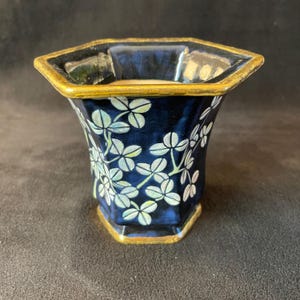 Puede incluir: Un jarrón hexagonal azul oscuro con borde y base dorados. El jarrón está decorado con un estampado floral blanco y verde claro. El jarrón tiene una forma y un diseño únicos.