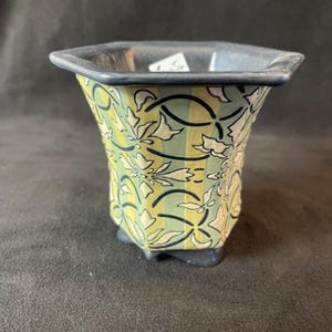 May include: A small ceramic vase with a square top and a flared body. The vase has a repeating floral pattern in white and black against a yellow and green striped background. The rim and base are dark blue.