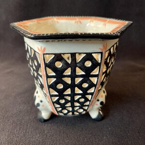 May include: A ceramic planter with a geometric black and white pattern. The planter has a square shape with a black rim and small feet. The interior is cream-coloured, and the exterior features cut-out designs.