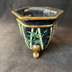 Puede incluir: Un pequeño cuenco decorativo negro y dorado con una forma única. El cuenco presenta un diseño de bambú con hojas verdes y rayas blancas. El borde y el pie son de color dorado.