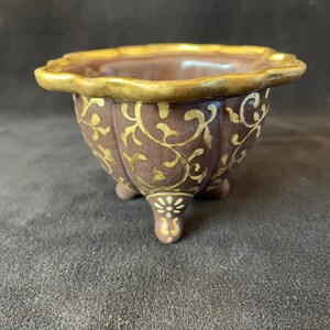 Può includere: Una ciotola in ceramica marrone a tre gambe con un bordo dorato smerlato. La ciotola è decorata con volute floreali dorate. La base di ogni gamba presenta un piccolo motivo floreale bianco.