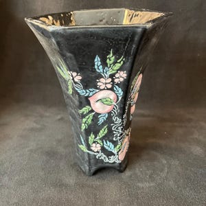 May include: Black ceramic vase with a flared, geometric shape. The vase is decorated with hand-painted pink peaches, green leaves, and small pink flowers. The vase has a square opening and sits on three small feet.