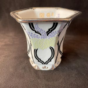 Könnte beinhalten: Eine weiße Keramikvase mit silbernem Rand und Boden. Die Vase hat ein Blumendesign mit Lavendel- und grünen Akzenten sowie schwarzen geschwungenen Linien. Der obere Rand hat goldene Buchstaben. Die Vase hat eine sechseckige Form.