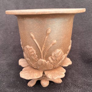 Könnte beinhalten: Ein brauner Keramik-Blumentopf mit breitem Rand und floralem Design. Der Topf wird von einem blütenförmigen Sockel mit einer Schildkröte am Boden getragen. Der Topf hat eine rustikale, erdige Ästhetik.