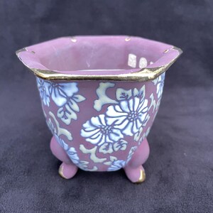 Puede incluir: Un pequeño cuenco decorativo de cerámica con exterior morado y borde dorado. El cuenco presenta un estampado floral en blanco y azul. El cuenco tiene tres pequeños pies con detalles dorados.