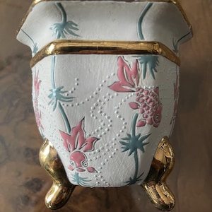 May include: A white ceramic planter with a gold rim and feet. The planter is decorated with pink fish and blue aquatic plants. The planter has a textured surface with small, raised dots. The gold feet are curved and add a touch of elegance.