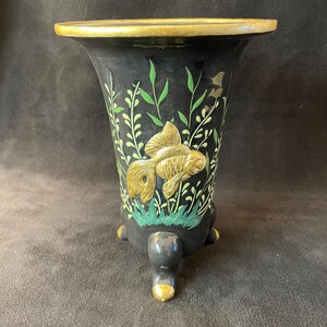 May include: A black ceramic vase with a flared rim and three rounded feet. The exterior features a gold-painted goldfish relief, green painted aquatic plants, and a gold-painted rim and feet tips. The vase has a smooth, dark finish.