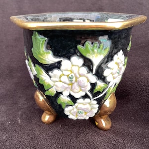 Puede incluir: Una pequeña maceta de cerámica negra con borde dorado y tres patas doradas. La maceta está decorada con diseños florales blancos y hojas verdes. Las flores tienen centros amarillos. La maceta es probablemente para plantas pequeñas o suculentas.