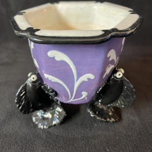 Puede incluir: Un cuenco decorativo de cerámica con exterior morado, detalles florales blancos y ribete negro. El cuenco está sostenido por dos patas negras estilizadas con forma de pez y aletas iridiscentes. El interior es blanco.