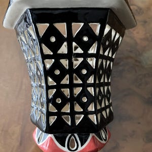 Könnte beinhalten: Eine schwarz-weiße dekorative Keramikvase mit geometrischem Ausschnittmuster. Die Vase hat einen rot-schwarzen Sockel und einen weißen Rand. Das geometrische Muster umfasst Dreiecke, Rauten und Kreise.