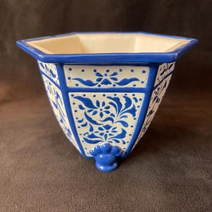 Puede incluir: Una maceta de cerámica azul y blanca con forma hexagonal. La maceta tiene un borde azul y está decorada con un estampado floral. La maceta tiene un pequeño pie azul en la parte inferior.