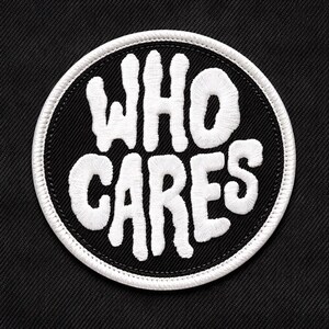 May include: A round, black patch with the embroidered words "WHO CARES" in white, encircled by a white border. The patch is set against a black fabric background, creating a high-contrast design.
