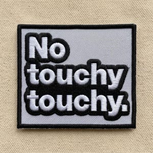May include: Square patch with the words "No touchy touchy" in white, outlined in black, on a light gray background. The patch has a black border and is likely an iron-on or sew-on patch.
