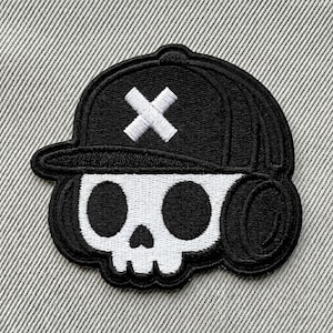 May include: Black and white embroidered patch featuring a skull wearing a baseball cap. The cap is black with a white cross on the front. The skull has large black eye sockets and a small nose and mouth.