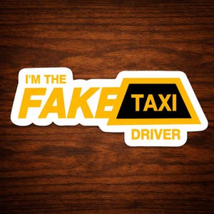 May include: A white sticker with the text "I'M THE FAKE TAXI DRIVER" in yellow and black. The sticker has a taxi cab design with a black top and yellow accents. The sticker is on a brown wooden surface.
