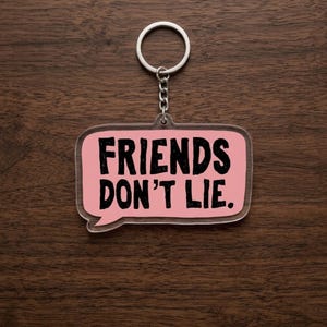 May include: A clear acrylic keychain with a pink speech bubble design. The keychain features the phrase "FRIENDS DON'T LIE" in bold black letters. The keychain is attached to a silver metal ring and chain, set against a dark wood grain background.