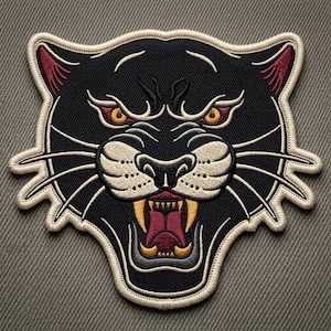 May include: Embroidered patch featuring a black panther with an open mouth, displaying sharp teeth. The panther has yellow eyes, red accents on the ears, and a cream-colored outline. The patch is designed in a traditional tattoo style.