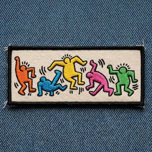 May include: Embroidered patch featuring a design of stylized figures in orange, blue, yellow, pink, and green. The figures are dancing on a white background with a black border. The patch is rectangular and appears to be sewn onto a denim fabric.