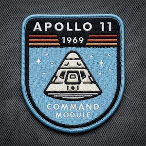May include: A blue, black, and white Apollo 11 Command Module patch. The patch features the text "APOLLO 11" above "1969" and a triangular spacecraft design. The words "COMMAND MODULE" are at the bottom.