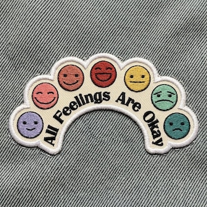 May include: Embroidered patch with a cream-colored border and the text "All Feelings Are Okay." The patch features seven colorful emoji faces, each expressing a different emotion, including happy, sad, and neutral.