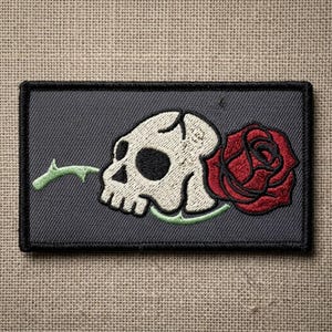 May include: Embroidered patch featuring a white skull with black eye sockets and a red rose. The skull is on a gray background with a black border. A green stem extends from the rose to the skull.