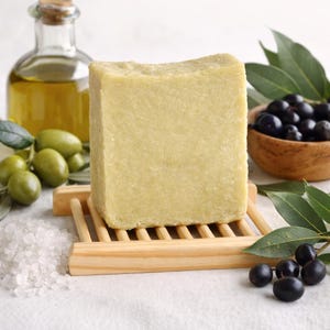 Può includere: Una saponetta verde chiaro è appoggiata su un portasapone in legno, circondata da olive, olio d'oliva in una bottiglia di vetro e una piccola ciotola di legno con olive nere. Sale marino bianco sparso nelle vicinanze. La scena evoca un'estetica naturale e da spa.
