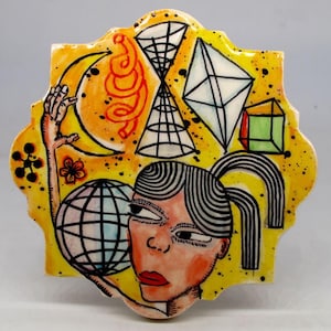 May include: A ceramic tile with a yellow background and a whimsical design. The artwork features a woman's face, a globe, geometric shapes, a hand reaching for a crescent moon, and other abstract elements. The tile has a decorative, scalloped edge.