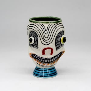 May include: A ceramic mug shaped like a stylized face with a green rim, black and white dotted patterns, large eyes, a red nose, and a wide toothy grin. The base features horizontal blue stripes, and the overall design includes intricate black line work.