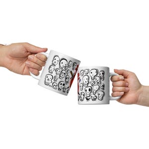 Può includere: Due tazze in ceramica bianca con illustrazioni di teschi neri. Una tazza presenta una grafica di mazza da baseball. Le tazze sono tenute su uno sfondo bianco. Un'idea regalo originale.