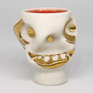 May include: A white ceramic skull-shaped mug with gold accents. The mug features a wide, toothy grin, eye sockets, and gold detailing around the eyes and cheekbones. The interior of the mug is red.