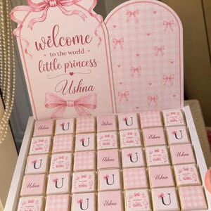 Peut inclure: Une boîte blanche contenant trente-cinq chocolats carrés emballés dans du papier vichy rose et blanc ou blanc avec le texte Ushna, U ou Mubarak Girl. La boîte présente un grand panneau rose avec des nœuds et le texte Welcome to the world little princess Ushna.