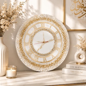 May include: A round wall clock featuring a white center dial with gold Roman numerals, surrounded by a textured border of gold and clear crushed glass accents. The clock has ornate gold hands and a thin red second hand, creating a layered design.