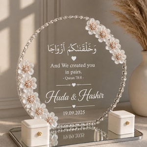 May include: A circular clear acrylic wedding sign featuring white floral accents, crystal-like trim, and white text reading "And We created you in pairs. - Quran 78:8 - Huda & Hashir 19.09.2025" with two small white ring boxes on a mirrored base.