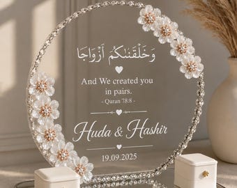 May include: A circular clear acrylic sign with a crystal-beaded rim, decorated with white floral accents. It features the Arabic text "وَخَلَقْنَاكُمْ أَزْوَاجًا", the English text "And We created you in pairs. - Quran 78:8 -", "Huda & Hashir", and "19.09.2025". Two small white ring boxes sit on the mirrored base.