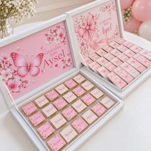 Peut inclure: Deux boîtes rectangulaires blanches remplies de petits chocolats carrés, chacun enveloppé dans un papier rose et blanc imprimé avec le nom Aysel. Les couvercles présentent des illustrations de papillons et de fleurs de cerisier.