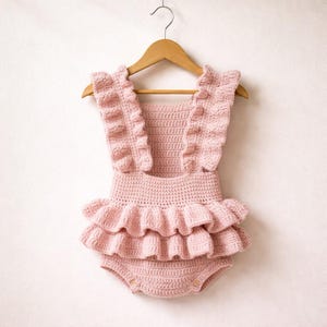 以下が含まれることがあります： フリル付きのストラップとウエスト周りのティアードフリルデザインが特徴の、淡いピンク色のクロシェ編みロンパース。ベビー服は、白い背景に木製のハンガーに展示されています。ロンパースにはボタン留めが付いています。