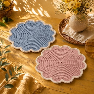 May include: Two round crochet doilies with scalloped edges, one in dusty blue and one in dusty pink, each featuring a cream-colored border. The circular mats are crafted with a dense, textured stitch pattern and are displayed on a wooden surface.