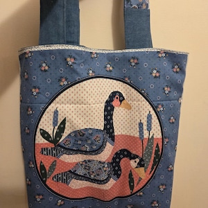 May include: A blue tote bag with denim handles and floral accents. The bag features a circular design with two geese in a pond scene. The background is a light blue fabric with small floral patterns.