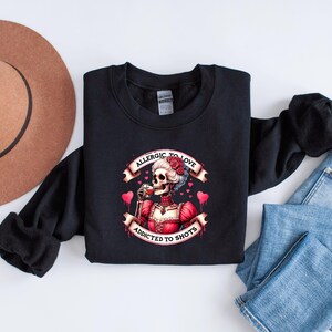 May include: Black sweatshirt featuring a graphic of a skeleton in a red dress, drinking from a glass. The text reads "Allergic to Love, Addicted to Shots" with heart accents. The sweatshirt is on a white surface.