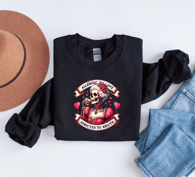 May include: Black sweatshirt featuring a graphic of a skeleton in a red dress, drinking from a glass. The text reads "Allergic to Love, Addicted to Shots" with heart accents. The sweatshirt is on a white surface.