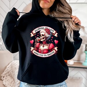 May include: Black hoodie featuring a graphic of a skeleton in a red dress, holding a drink, with the text "ALLERGIC TO LOVE ADDICTED TO SHOTS". The design includes hearts and roses.
