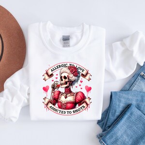 May include: White sweatshirt with a graphic of a skeleton in a red dress, drinking from a glass. The text on the banner reads "Allergic to Love, Addicted to Shots." The design includes red hearts and roses.