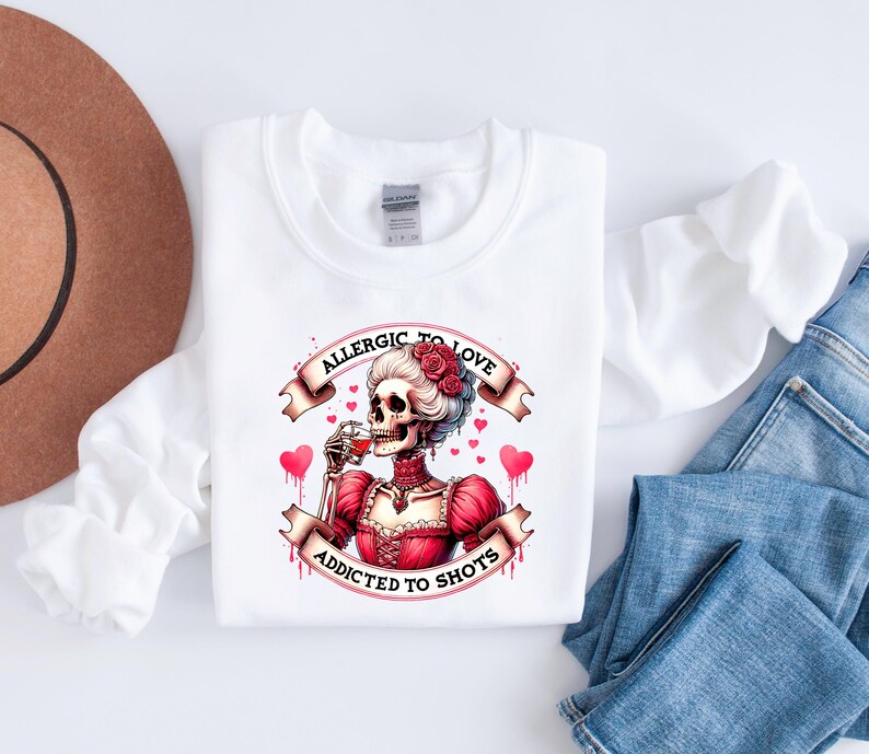 May include: White sweatshirt with a graphic of a skeleton in a red dress, drinking from a glass. The text on the banner reads "Allergic to Love, Addicted to Shots." The design includes red hearts and roses.